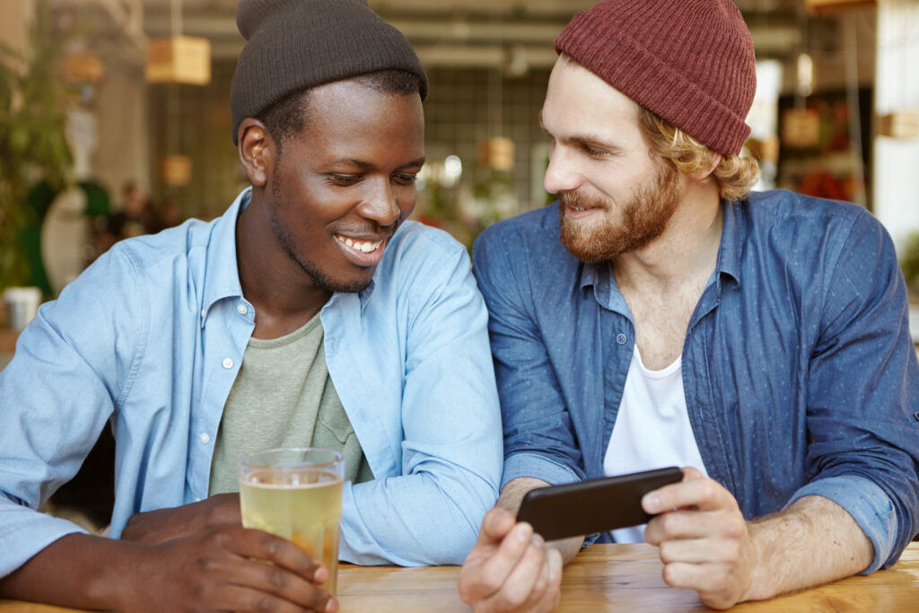 Two men in a coffee shop. One shows the other something on his phone, they are both smiling